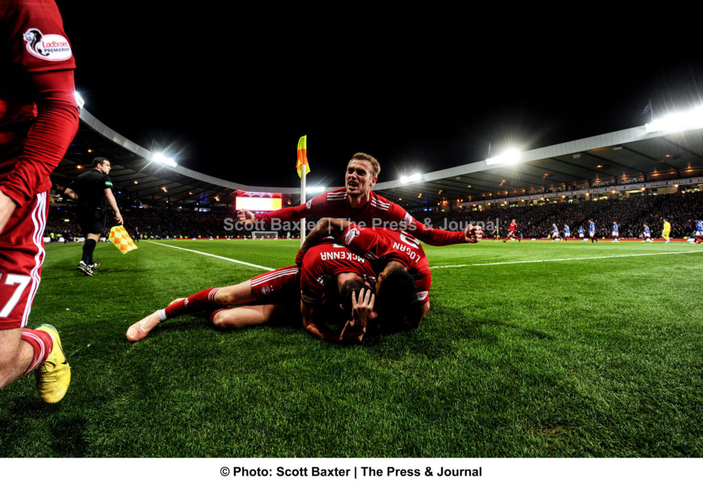 A wide angle photograph of players on top of one another celebrating during a football match between Aberdeen v Rangers. Aberdeen win 1-0.