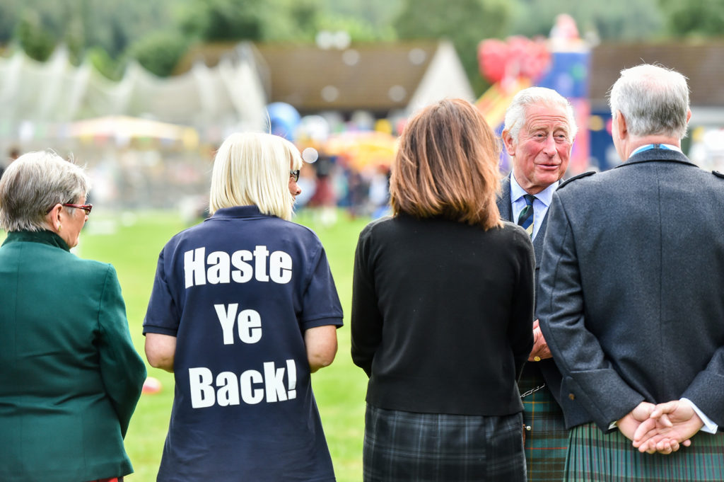 His Royal Highness The Prince of Wales greets people at the Ballater Highland Games. The four people a photographed from behind with Prince Phillip looking towards the camera. One person has written on the back of their black t-shirt Haste Ye Back.