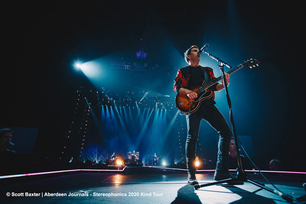 Press Photography by Scott Cameron Baxter. Image show Stereophonics lead singer performing on stage at microphone. Blue lights shine all around him. Picture taken with a wide angle lens at the height of lead singers foot at base end of stage.