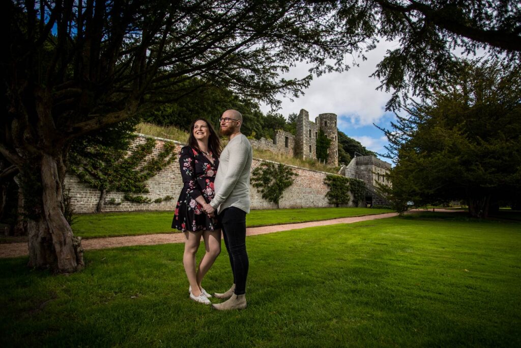 A couple pose in Ellon Castle Gardens for an engagement photoshoot. A sunny day and the couple are photographed under the cover of overhanging trees with the remains o the castle over their right shoulders in the background. The couple are facing one another while holding hands. She wears a dress floral dress and he wears a grey jumper with dark jeans.