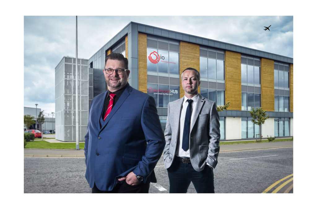 Two men in suits, one blue and one gray pose in front of their glass office building for a photograph on a cloudy day. PR and Commercial photography by Scott Cameron Baxter Aberdeen.