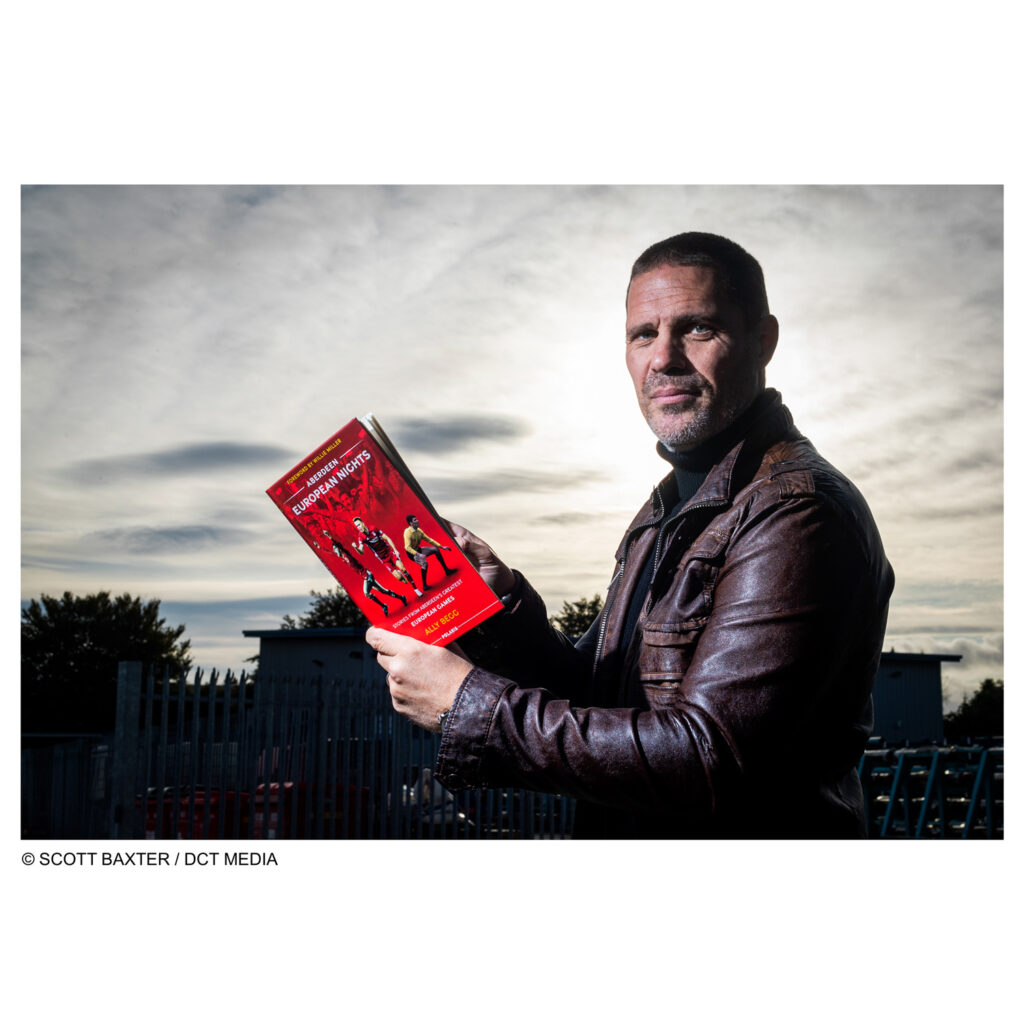 Picture shows Ally Begg, author, holding his book while looking at the camera, the sky is dramatic with clouds. The sun is directly behind his head. 