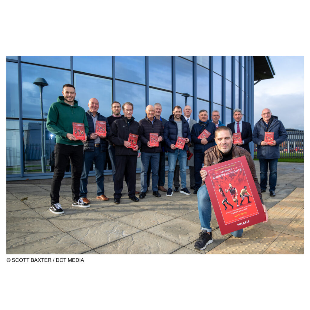 Picture shows Ally berg, Author, with 12 former and current footballers of Aberdeen Football Club promoting his new book launch. He holds a large copy of the front of his book while the players stand behind him with copies of the book.