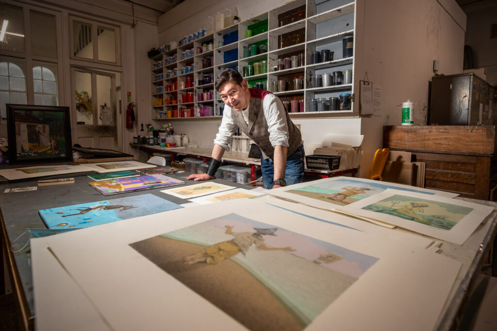 Charlie Hynes at a large table covered in his prints. He wears a white shirt with a tweed waistcoat, in a printers studio. there are lots of paint and art materials all around.