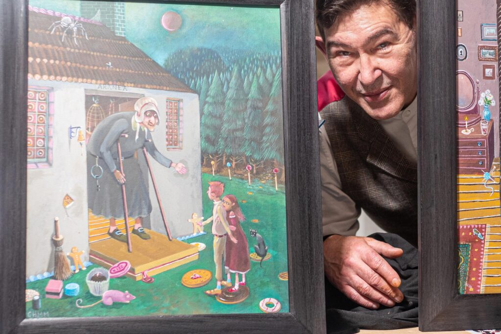 Screenprinter Charlie Hynes, poses for a photograph with his artwork. he is seen here ducking down to be level with a painting which shows an old woman and children.