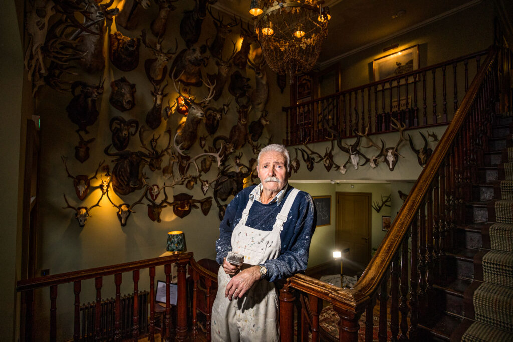 A decorator stands on a staircase with a paintbrush in hand. behind him, there are trophy hunt deer antlers displayed completely covering the wall, maybe 100 trophies. The man wears a paint covered white apron with a blue jumper.
