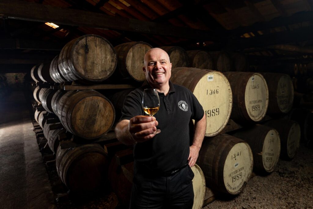 A distillery manager stands with a glass of scotch whisky in the barrel warehouse, raising a glass to the camera smiling. He is wearing all black and the wooden barrels are being him.