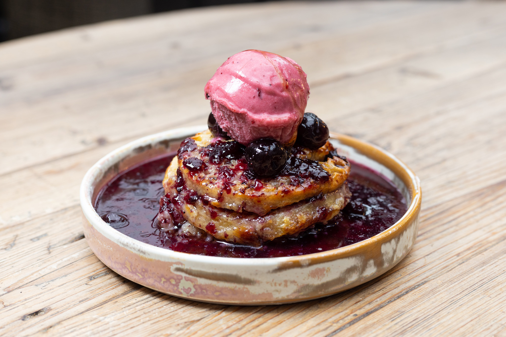 Side on photograph of a crumpet and  strawberry sorbet food dessert, plate is on a wooden table in a beige coloured plate. with black cherries topped with the sorbet. Food photography Aberdeen by Scott Cameron Baxter