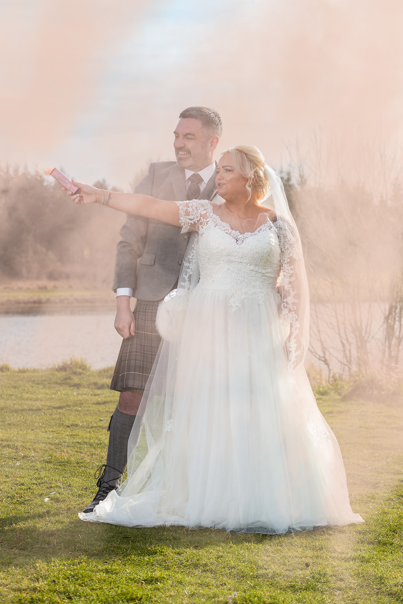 Colour image of a couple letting off a red flare, on the banks of a loch. The bride is at the front centre while the groom stands directly behind her, looking at her extended arm holding the red flare. both are happy.Wedding photographer Aberdeen, Picture by Scott Cameron Baxter.