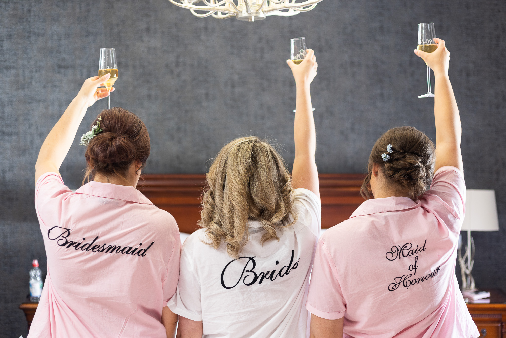 A bridal party hold up drinks with their backs turned away, on the back of their pyjamas it reads Bridesmaid (Pink pyjamas), Bride (White pyjamas), Maid of honour (Pink pyjamas). They are in a hotel room hours before the ceremony.