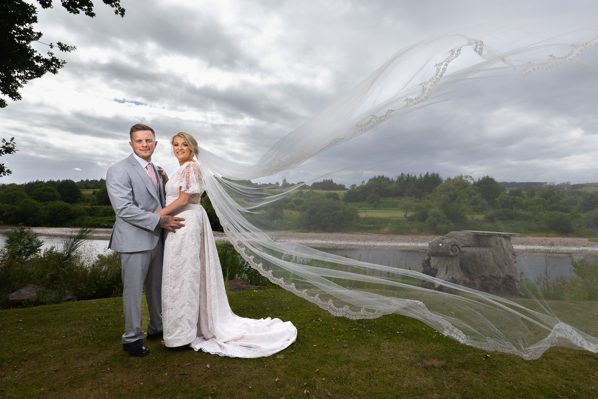 A colour image of a couple who are posing for a picture on the banks of the River Dee at Maryculter House Hotel. The couple are at the left of the picture holding onto one another waists. He is wearing a light blue suit, she is wearing a white wedding dress with a long veil which has blown to the right of the image. Wedding photographer Aberdeen, Picture by Scott Cameron Baxter.