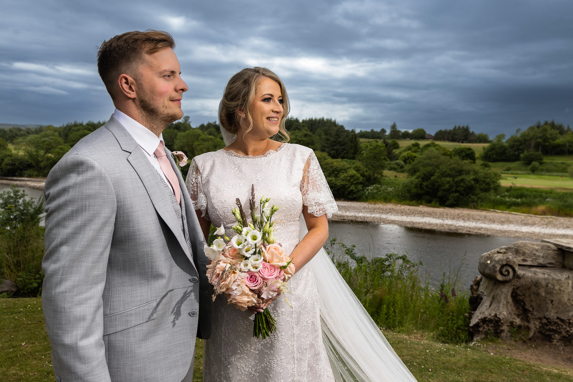 A colour image of a couple who are posing for a picture on the banks of the River Dee at Maryculter House Hotel. The couple are at the left of the picture holding onto one another waists. He is wearing a light blue suit, she is wearing a white wedding dress. They are looking to the right of the image. Wedding photographer Aberdeen, Picture by Scott Cameron Baxter.
