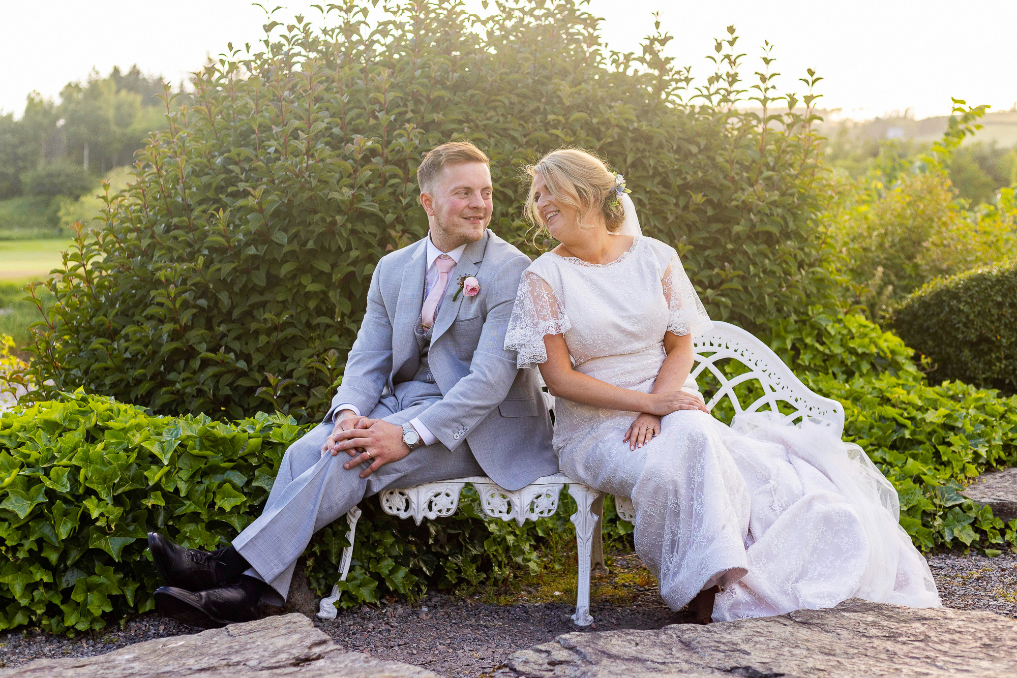 A colour image of a couple who are sitting on a white metal bench, in the centre of the image after having been married. He is on the left wearing a blue suit, she is wearing a white long wedding dress, the sun is casting a warm glow around the green trees and bushes behind them. Wedding photographer Aberdeen, Picture by Scott Cameron Baxter.