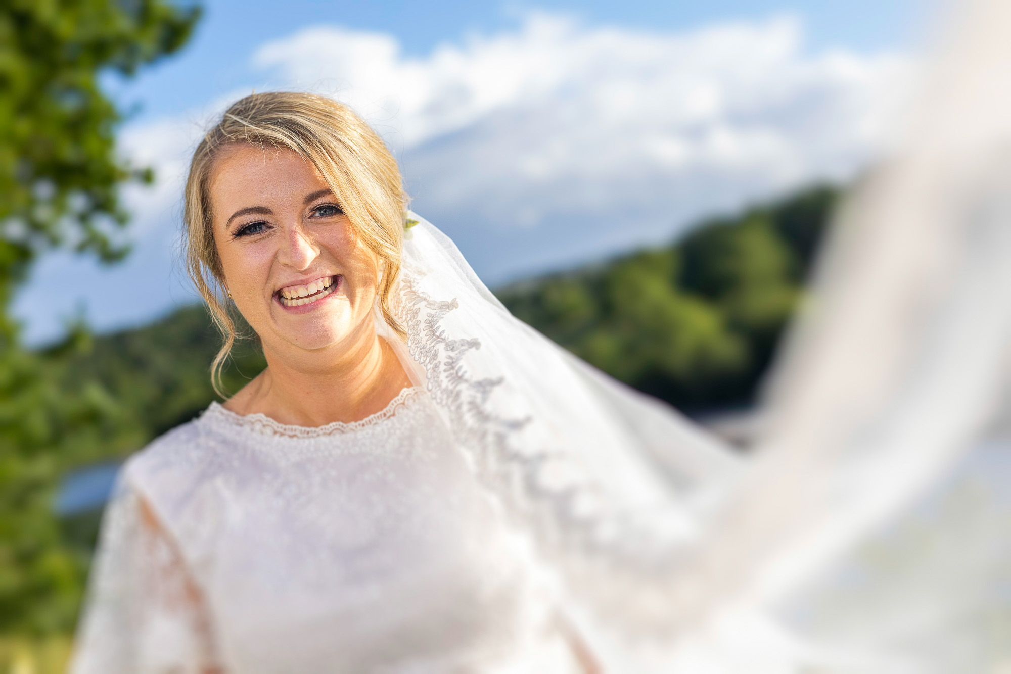 A colour image of a bride smiling directly into the camera. There is a blue sky, green trees, the image is taken at a slight tilt and the brides veil come over towards the lens whith the bride off the the left of the image. The surroundings are blurred with the brides face pin sharp and happy in expression. . Wedding photographer Aberdeen, Picture by Scott Cameron Baxter.