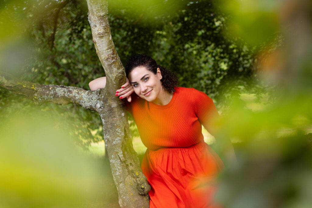 PR Photo of a woman wearing all red dress, sitting on a tree branch, the outside of the frame has green leaf all around while she in directly in the centre of the frame with her head against another branch resting on her hand. Image by Aberdeen Press photographer Scott Cameron Baxter.