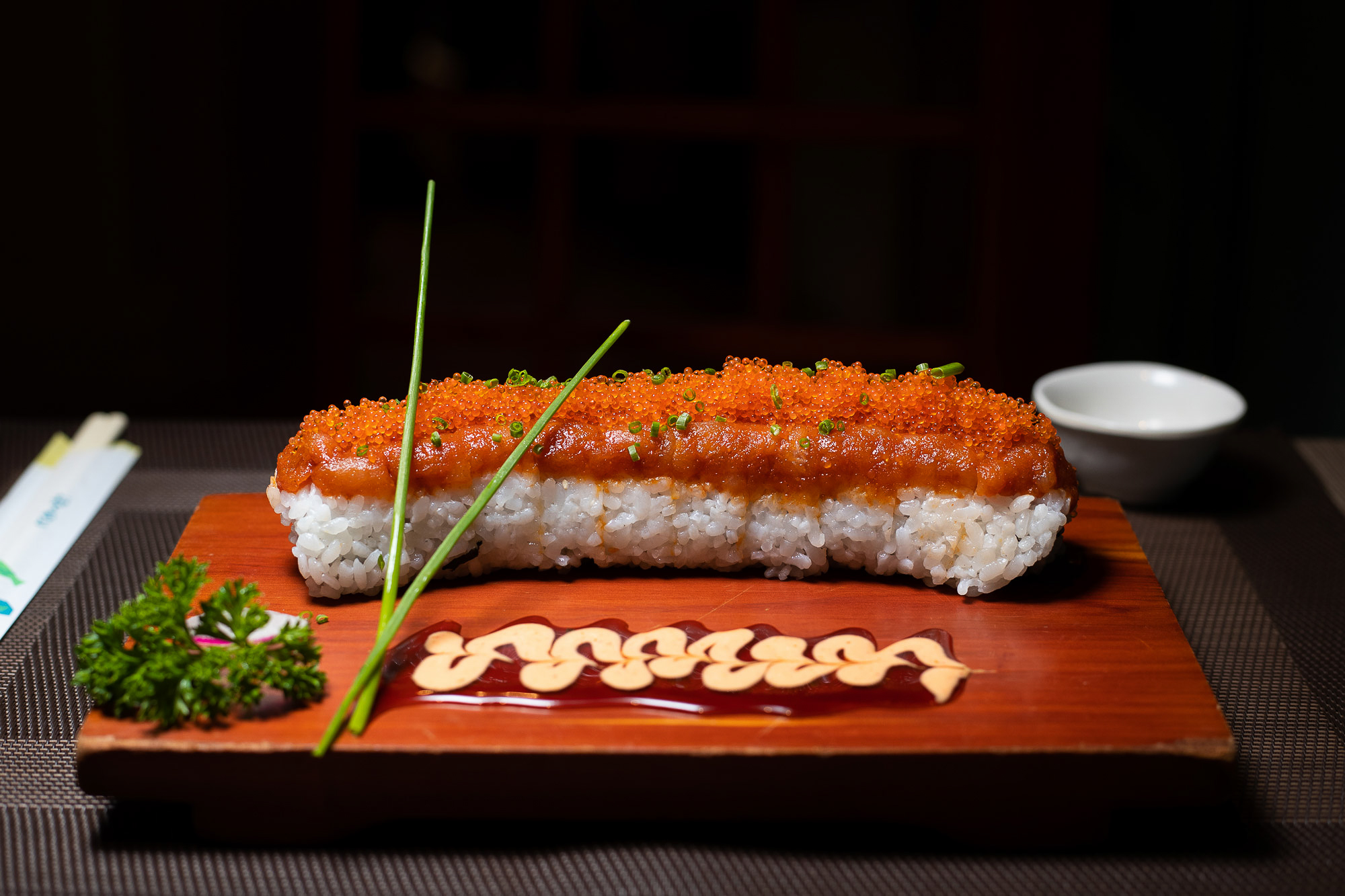 Sushi roll with fish eggs and salmon presented on a dark red wooden board. With a spicy red and yellow sauce with a wavy pattern. Dish is framed in centre of image with a black background. Aberdeen Food photographer Scott Cameron Baxter.