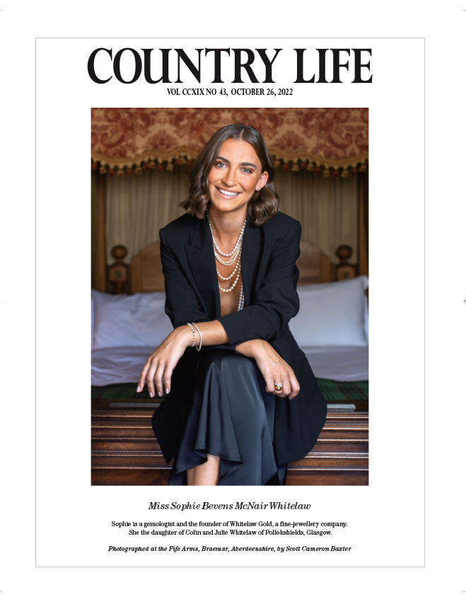 Front cover of Country Life Magazine, Shows a woman at the end of a bed in the Fife Arms Hotel, Ballater, Aberdeenshire. She is wearing a black blazer and dark skirt. Photographed by Scott Cameron Baxter for Country Life Magazine.