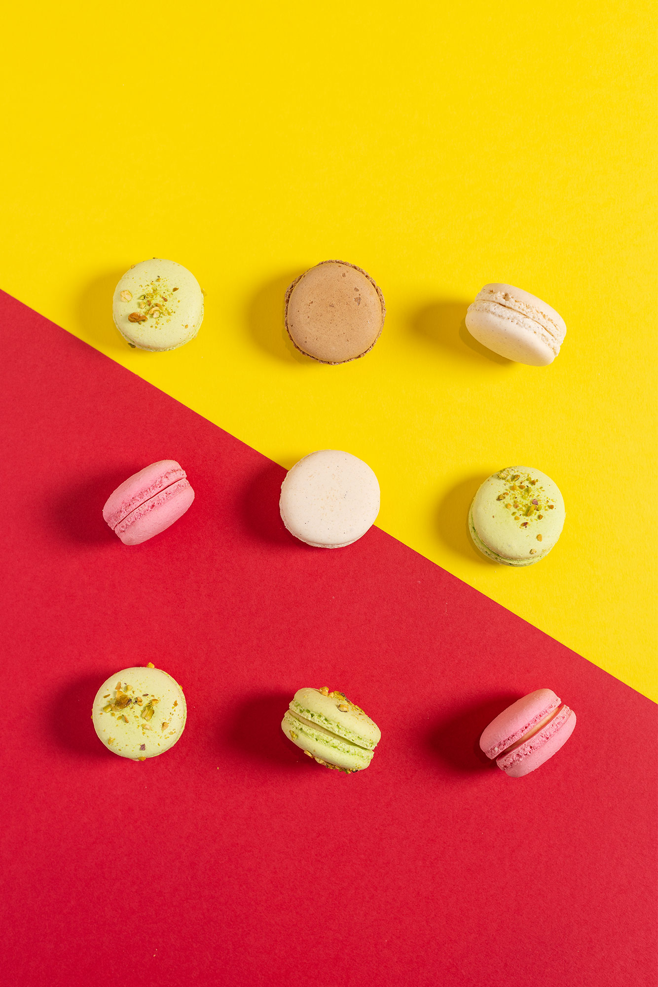 Food photographer Scott Cameron Baxter. Pictures shows 9 macarons from a birdseye view on a half yellow and red table. The colour difference is at a 45 degree angle, yellow at the top, red at the bottom. with the centre macaron splitting the two colours.