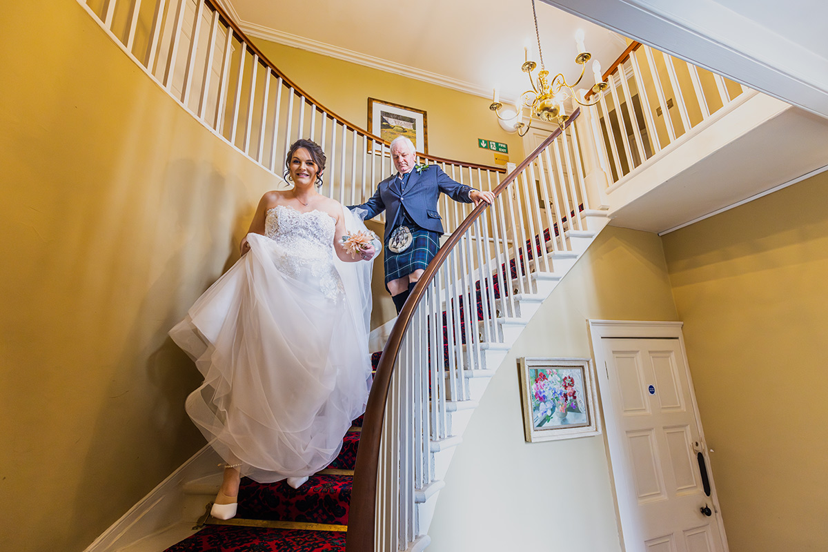 A colour image of a bride and her father walking down a curved set of stairs. The railing is wooden and the struts are white, carpet red. They are walking toward the camera.. Wedding photographer Aberdeen, Picture by Scott Cameron Baxter.