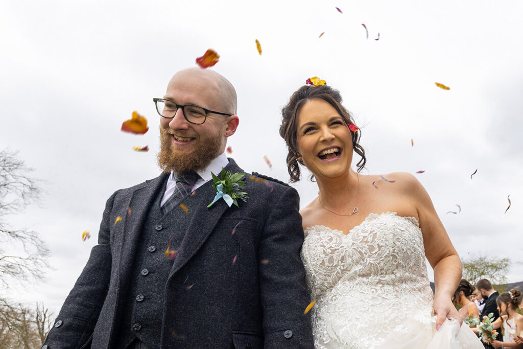 A couple have just been married at a scottish wedding. Groom on the righjt and bride on the left smiling just above the camera, confetti has been thrown over them both. The background is clouds. He wears a tweed jacket and tartan tie and she is wearing a white wedding dress. Picture taken by Wedding Photographer Aberdeen by Scott Cameron Baxter