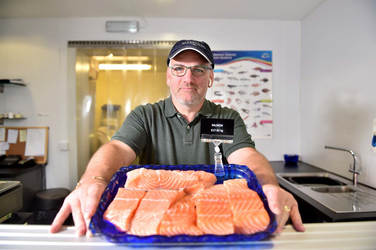 Aberdeen Food and drink photographer Scott Cameron Baxter. Picture shows a fishmonger holding a blue tray of fresh pink salmon on a countertop. The fishmonger is a man wearing a green polo shirt and a dark hat in his shop. He iand the tray of fish are lit from the right, he and the tray are in the middle of the photograph.