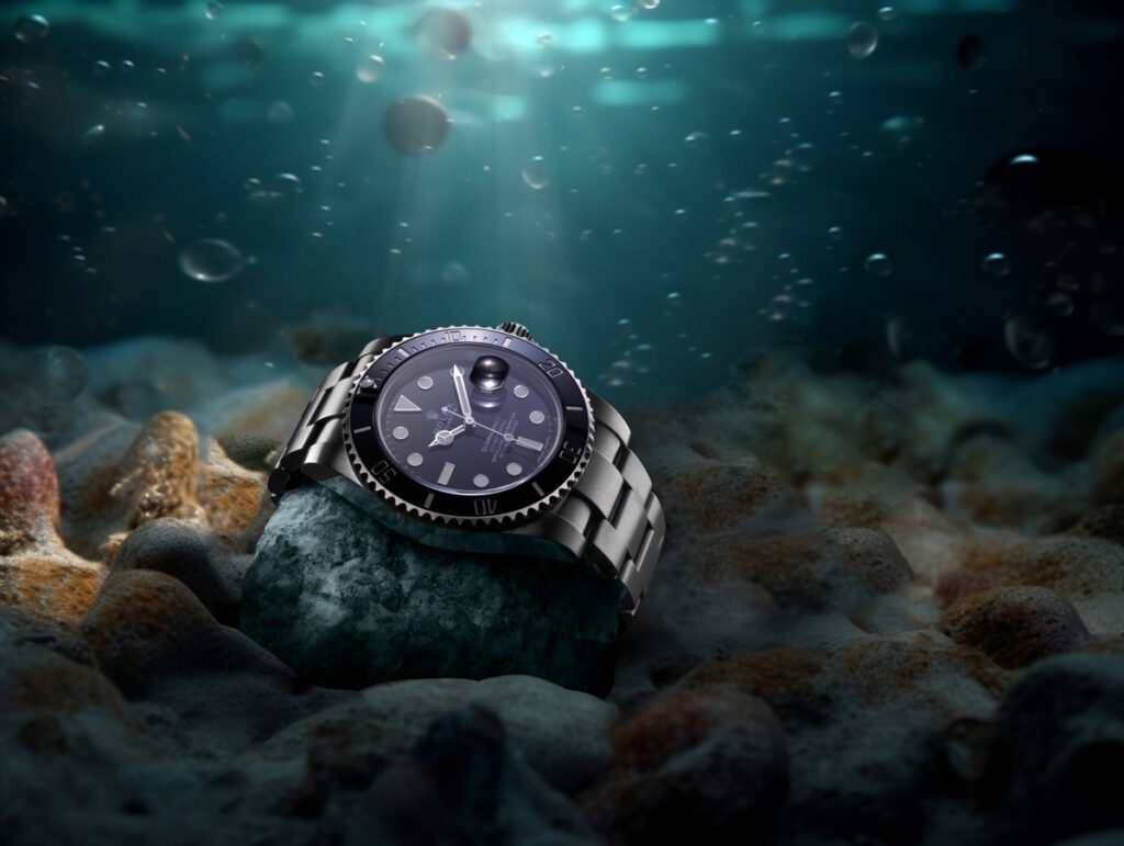 Commercial Product Photographer Aberdeen - Scott Cameron Baxter. Image shows a Rolex Submariner watch underwater sitting on a rock on the seabed. Light is coming from behind above on the surface which lights the watch and its face. It is mainly aqua blue with the watch being silver. Commercial photography Aberdeen.