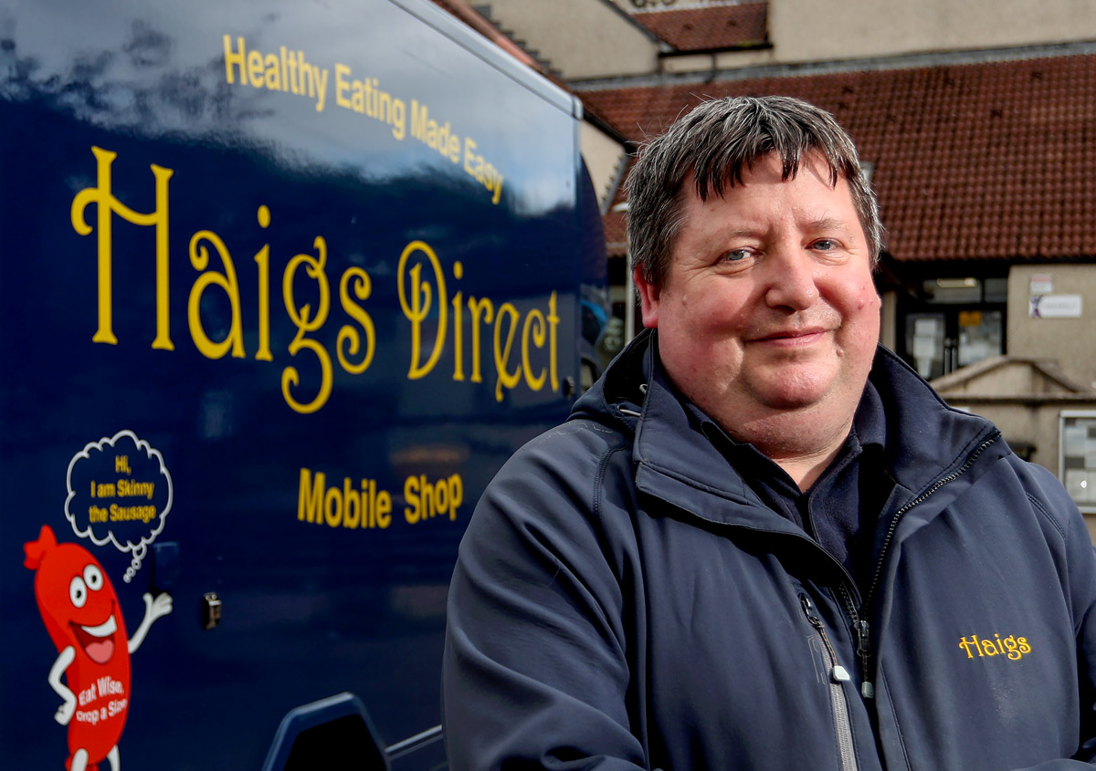 Aberdeen Food and drink photographer Scott Cameron Baxter. Picture shows a man from HAIGS BUTCHER in Aberdeen, standing outside in front of his portable butcher produce van, which is navy and has in yellow letters HAIGS DIRECT on the side. The man is on the right of the image and the van on the left, the image is tightly cropped to show only the man and the writing on the side of the van.