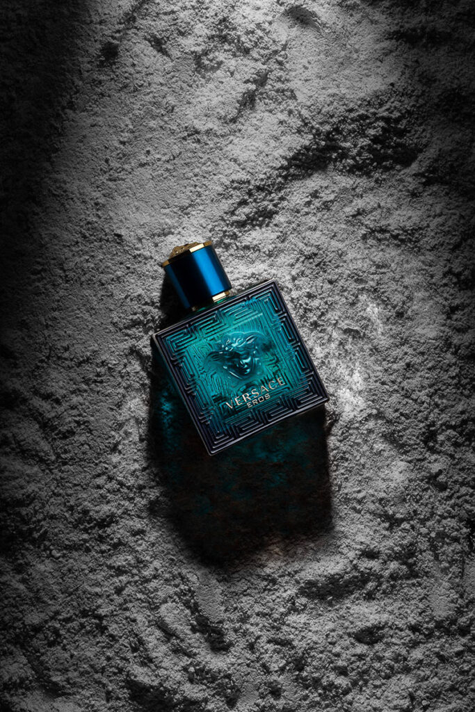 Commercial Scotland Product Photographer Aberdeen - Scott Cameron Baxter. Image shows a aqua blue aftershave bottle of Versace EROS. It is laying flat, at an angle with the top of the bottle at 10 o'clock. the square bottle is laying on a fine powder which looks like white sand. The bottle is dramatically lit from the top and behind which makes the bottle stand out against the white sand.