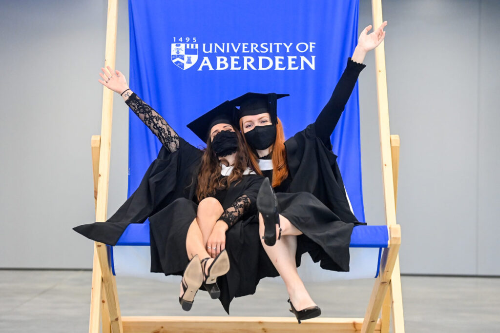 Two graduates celebrating their graduation wearing black gowns and graduation caps. Sitting on a blue oversized University of Aberdeen deck chair.