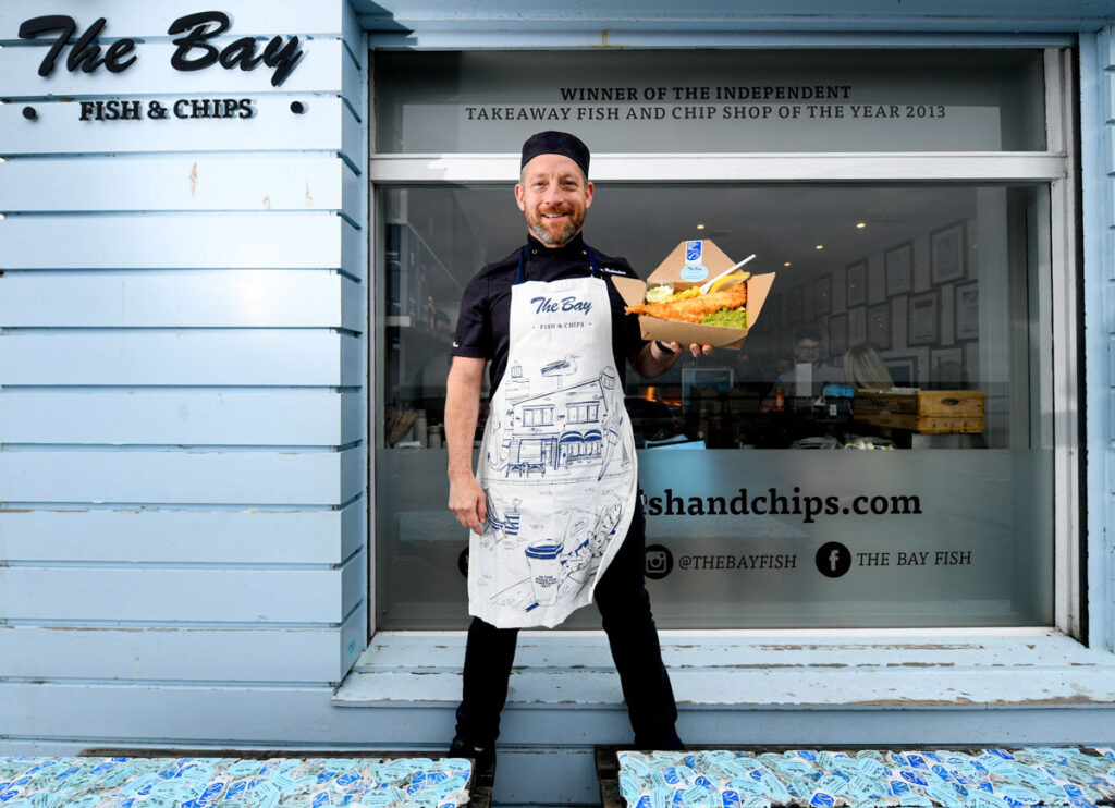 The owner of The Bay Fish & Chips in Stonehaven stands in front of the restaurant window holding a portion of their famous fish and chips. He is wearing an apron adorned with the company logo.