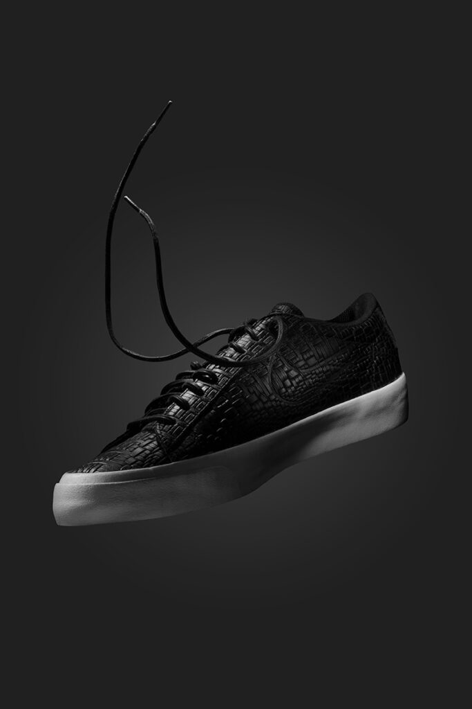 A picture of a Black and White Nike trainer. 
 The shoe is suspended in mid air and the laces are floating above. The background is dark and grey, lighter in the middle and darker on the outside. Photographed by commercial product photographer Scott Cameron Baxter from Aberdeen, Scotland.