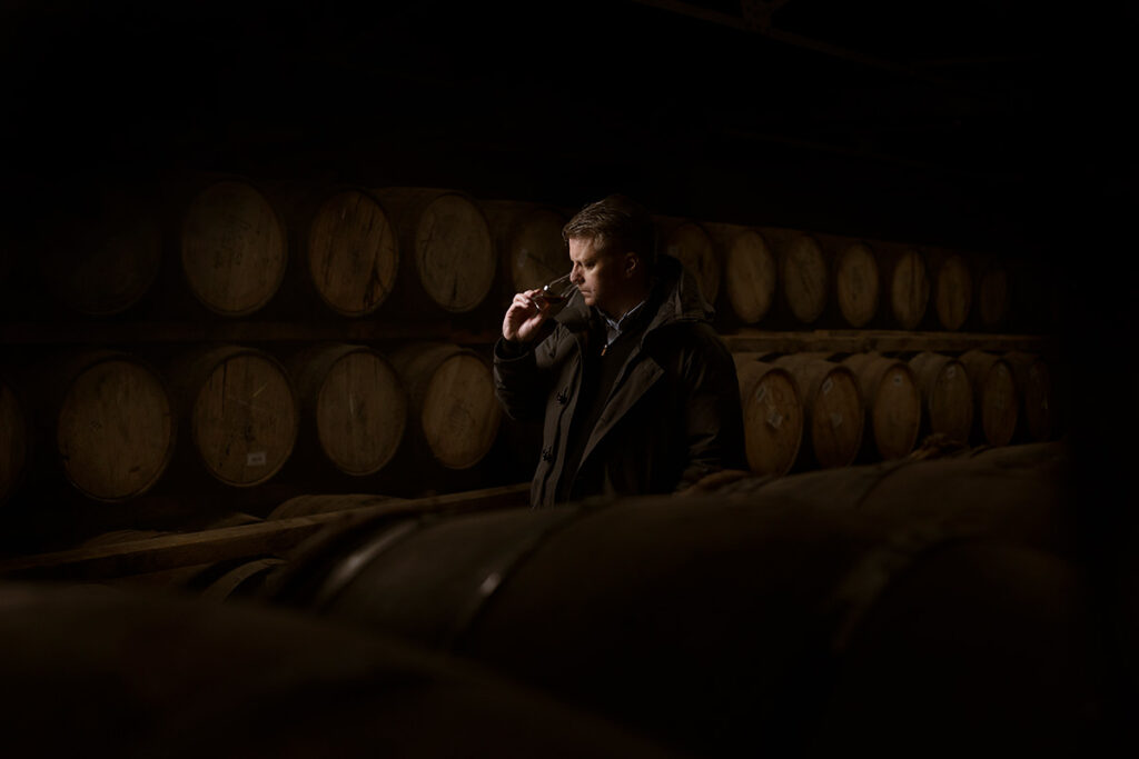 Brian Kinsman, master distiller for William Grant & Sons, including brands like Glenfiddich, is Photographed by Scottish product and commercial photographer Scott Cameron Baxter, Aberdeen Scotland. Brian, wearing a black jacket stands amongst hundreds of casks smelling and tasting whisky from a Glencairn's glass. He is dramatically lit in this dark but warm and moody shot.