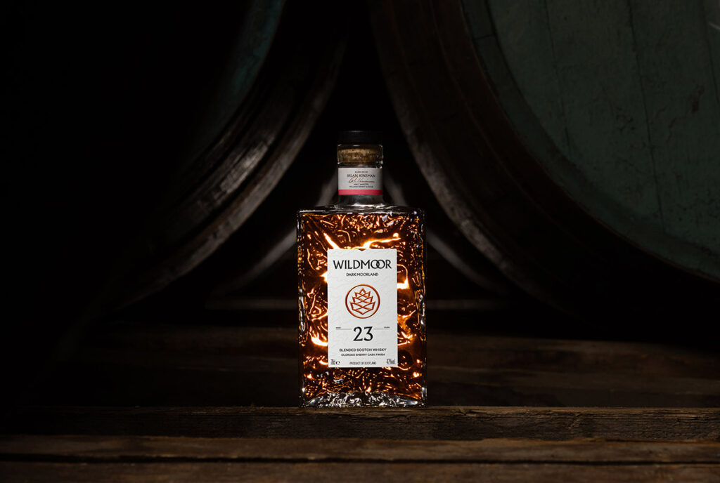 A Wildmoor Scotch Whisky bottle sits between two casks on a wooden surface, the bottle is lit from behind giving off a bright warm glow. The bottle is rectangular in shape and is contoured on its glass surface. Photographed by Scottish product and commercial photographer Scott Cameron Baxter, Aberdeen Scotland.