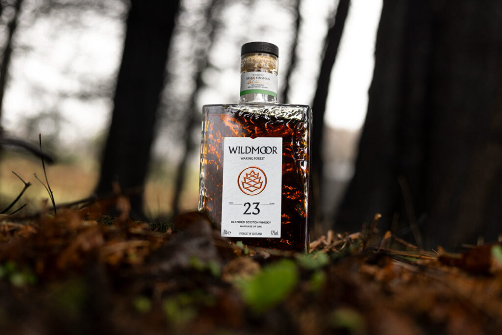 A Wildmoor Waking Forest 23 year old Scotch Whisky bottle stands on an autumn surface of leaves and twigs. The bottle is outside and lit from behind with natural light. The bottle is rectangular in shape and is contoured on its glass surface. The foreground is slightly obscured with leaves and the background is trees. Photographed by Scottish product and commercial photographer Scott Cameron Baxter, Aberdeen Scotland.